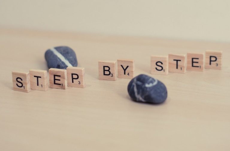 Wooden letters spelling "STEP BY STEP" next to smooth stones on a light surface.